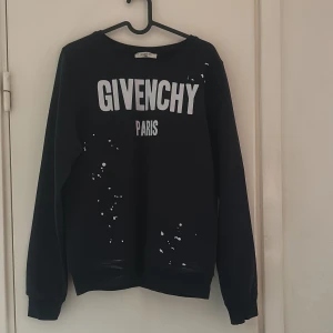Svart sweatshirt från Givenchy Paris - Svart sweatshirt från Givenchy Paris med vit logotyptryck framtill och små vita stänkdetaljer. Klassisk rund halsringning och långa ärmar. Perfekt för en trendig och avslappnad look.