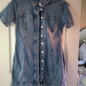 Jeansklänning med knappar från Zara - Säljer en snygg jeansklänning från Zara i storlek L. Klänningen har korta ärmar, två bröstfickor och knäppning hela vägen framtill. Perfekt för dig som gillar denim och vill ha en enkel men trendig look.