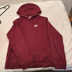 Vinröd hoodie från Nike - Säljer en vinröd hoodie från Nike med broderad vit logga på bröstet. Tröjan har huva och en stor magficka framtill. Perfekt för dig som gillar en enkel och stilren look.