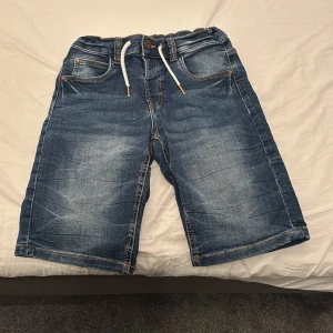 Jeansshorts med snörning från LAB Industries - Snygga blå jeansshorts från LAB Industries med vit snörning i midjan och klassiska fickor både fram och bak. Perfekta för sommaren och har en avslappnad passform. Tillverkade i denim för en cool look.