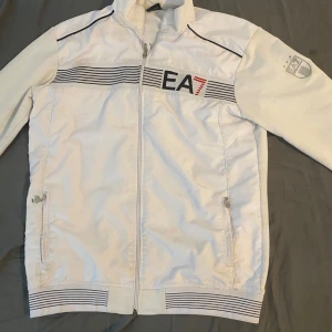 Vit jacka från Emporio Armani EA7 - Snygg vit jacka från Emporio Armani EA7 med dragkedja framtill och svarta detaljer. Jackan har ribbade muddar, två fickor med dragkedja och tryckt logga på bröstet samt ryggen. Perfekt för en sportig och stilren look.
