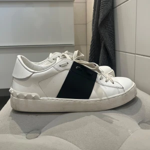 Valentino Garavani sneakers med svart detalj - Säljer ett par vita Valentino Garavani sneakers med en bred svart rem över sidan och ikoniska nitar på hälen. Perfekta för dig som gillar stilrena och lyxiga sneakers.Ser nästintill helt nya ut eftersom endast använt några få gånger. Passar superbra nu till student eller avslutningar och sitter som en smäck. Orginalpris 7000kr+ men säljer billigare på grund av använda. Men öppen för prutning❤️