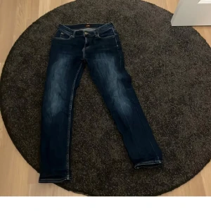 Mörkblå jeans från Lee - Säljer ett par mörkblå jeans från Lee med klassisk femficksdesign och raka ben. Jeansen har snygga slitningar framtill och tydliga sömmar. Passar dig som vill ha en tidlös och enkel stil.