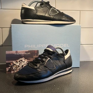 Svarta sneakers från Philippe Model Strl 41 - Säljer ett par svarta och blå Philippe Model sneakers med vita och blå detaljer. Skorna har snörning, rund tå och platt sula. Tungan på skon är mörkblå, men framstår som lila på bilden. Ovandelen är i en mix av skinn och textil, vilket ger en sportig men stilren look. Perfekta för dig som gillar klassiska men trendiga sneakers. Använt skick, men har mycket kvar att ge! Tveka inte att höra av er vid funderingar, eller om ni vill ha flera bilder!  👏🤝