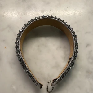 Glittrigt pärlprytt diadem - Säljer ett brett diadem dekorerat med glittriga, mörkgrå pärlor och små metallpärlor. Insidan är klädd i beige mockaliknande material. 