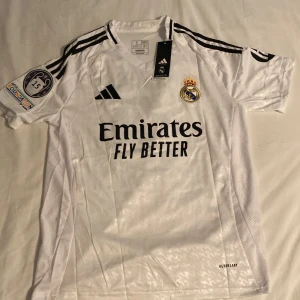 Real Madrid hemmatröja 2023/24 - Vit Real Madrid fotbollströja från Adidas med svarta detaljer och klubbmärke på bröstet. Tröjan har korta ärmar, broderat klubbemblem, Champions League-märke med siffran 15 och sponsorloggor. Perfekt för fans som vill visa sitt stöd.