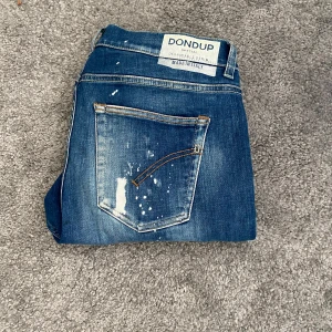 Dondup jeans - Säljer ett par blå jeans från Dondup med coola slitningar och färgstänk. Jeansen har klassisk femficksdesign och normal passform. Perfekta för dig som gillar en avslappnad och trendig look.