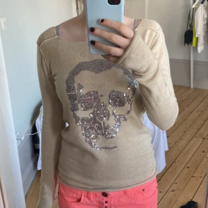 Beige tröja med paljett-dödskalle - Säljer en beige långärmad tröja med en stor dödskalle i silverfärgade paljetter på framsidan. Tröjan har en rund halsringning och är i ett mjukt material, perfekt för en cool och avslappnad stil.