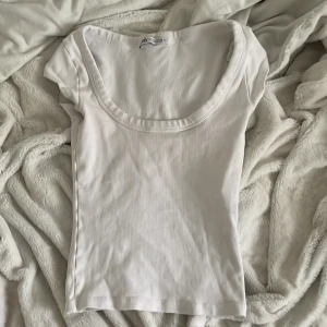 Vit ribbad topp från Zara - Säljer en vit ribbad topp från Zara med rund, djup urringning. Toppen har korta ärmar och är tillverkad i ett mjukt, stretchigt material som sitter tight mot kroppen. Perfekt att styla med jeans eller kjol. Nöstan oanvänd, var köpt i italien, skriv om ni har frågor 💖💞