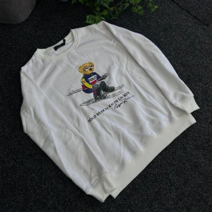 Vit sweatshirt med Polo Bear från Ralph Lauren - Vit sweatshirt från Ralph Lauren med ikoniskt Polo Bear-tryck på framsidan. Tröjan har rund halsringning och långa ärmar. Tillverkad i 100% bomull för en mjuk och bekväm känsla. Perfekt för dig som gillar klassisk och sportig stil.
