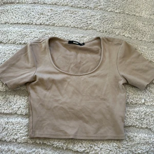 Beige croppad t-shirt från Boohoo - Säljer en beige croppad t-shirt från Boohoo med rundad, djup urringning och korta ärmar. Perfekt till sommaren och enkel att matcha med olika outfits.
