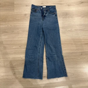 Blå wide jeans med råa kanter från H&M - Säljer ett par blå jeans med hög midja och vida ben. Jeansen har råa, avklippta benslut och klassiska fem fickor. Perfekta för en avslappnad och trendig look.