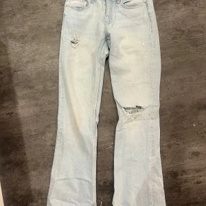Ljusblå bootcut jeans från Gina Tricot - Ljusblå jeans med bootcut passform från Gina Tricot. Byxorna har slitna detaljer vid knä och lår, klassisk femficksdesign och knappgylf. Perfekta för en avslappnad och trendig look.