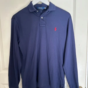Blå långärmad pikétröja från Polo Ralph Lauren - Säljer en klassisk blå långärmad pikétröja från Polo Ralph Lauren med röd broderad logga på bröstet. Tröjan har krage och knäppning framtill, samt en slim passform. Perfekt för en stilren look.