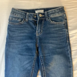 Blå bootcut jeans från Lindex - Säljer ett par klassiska blå jeans från Lindex med bootcut-modell. Jeansen har vita kontrastsömmar, fem fickor och snygga detaljer på bakfickorna. Storlek 140.