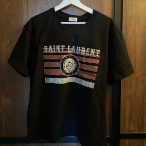 Svart t-shirt från Saint Laurent med tryck - Svart t-shirt från Saint Laurent med ett stort tryck framtill i vitt och rött. Trycket har texten 'SAINT LAURENT' och ett emblem i mitten. Klassisk rund hals och korta ärmar. Perfekt för en trendig och avslappnad stil.