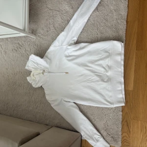 Vit hoodie från Moose knuckles med gulddetaljer - Säljer en stilren vit hoodie från Moose knuckles med unika guldfärgade detaljer på ena ärmen. Tröjan har huva med snörning och långa ärmar. Perfekt för dig som gillar en clean look med en twist.