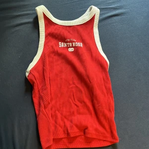 Röd ribbad linnetopp från H&M - Säljer en röd ribbad linnetopp från H&M med vita kanter och texten 'Santa Rosa' framtill. Toppen har ärmlös design och sportig look, perfekt för varma dagar eller träning.