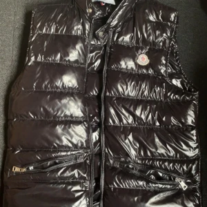 Svart dunväst från Moncler - Snygg svart dunväst från Moncler med glansig finish och klassisk logotyp på bröstet. Västen har två framfickor med dragkedja och quiltad design. Perfekt för lager-på-lager och stilrena outfits.