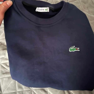 Mörkblå sweatshirt från Lacoste - Säljer en klassisk mörkblå sweatshirt från Lacoste med rund halsringning och den ikoniska krokodilloggan broderad på bröstet. Perfekt för en stilren och avslappnad look.