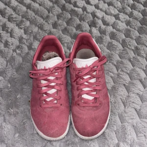 Adidas Gazelle rosa sneakers - Säljer ett par rosa Adidas Gazelle sneakers med vita detaljer och snörning. Skorna har klassisk låg profil, vit sula och är tillverkade i mocka. Perfekta för dig som gillar en sportig och stilren look.