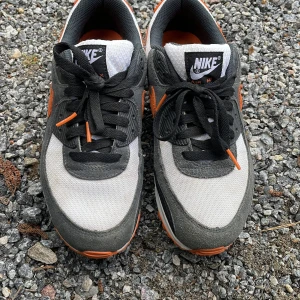 Nike Air Max 90 i grå, vit och orange - Säljer ett par Nike Air Max 90 med grå mockadetaljer, vit mesh och orange swoosh samt detaljer. Klassisk siluett med svart och orange på mellansulan och hälpartiet. Skorna har snörning och synlig Air-enhet i sulan.