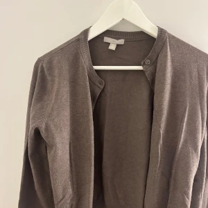 Brun kofta med rund hals - Säljer en enkel brun kofta med rund hals och långa ärmar. Koftan har ribbade muddar och knappstängning framtill. Perfekt att slänga över en topp för en avslappnad look.