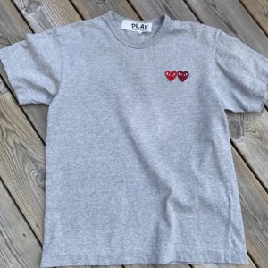 Grå t-shirt från Comme des Garçons Play - Säljer en grå t-shirt från Comme des Garçons Play med två röda hjärtan broderade på bröstet. Klassisk rund hals och korta ärmar. Perfekt basic med en twist.