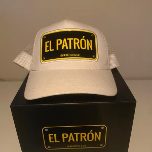 El Patrón keps från John Hatter & Co - Snygg keps från John Hatter & Co med texten 'El Patrón' på en skylt framtill. Finns i både vit och svart färg med gult tryck. Klassisk böjd skärm och mesh på sidorna för extra ventilation.