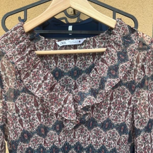 Blommig volangklänning från Zara - Supersöt klänning från Zara med volangdetaljer och lång ärm. Klänningen har ett mönster i blått, vinrött och beige, smock i midjan och volanger upptill och nertill. 