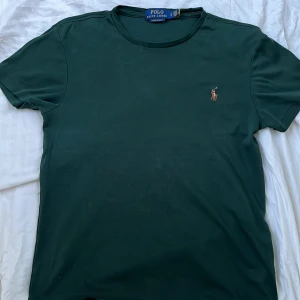 Mörkgrön t-shirt från Polo Ralph Lauren - Passar 13-14 åringar. Kostar 1200kr ny. Snygg mörkgrön t-shirt från Polo Ralph Lauren med klassisk broderad logga på bröstet. Modellen är slim fit och har rund halsringning samt korta ärmar. Perfekt för en stilren och enkel look.