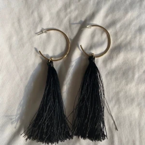 Svarta tofsörhängen med guldfärgade ringar - Säljer ett par stora örhängen med svarta tofsar och guldfärgade ringar. Perfekta för att ge en cool touch till din outfit. Lätta att bära och riktigt snygga statementörhängen.