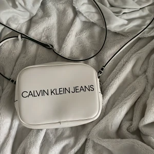 Vit axelväska från Calvin Klein Jeans - Säljer en stilren vit axelväska från Calvin Klein Jeans med svart logotyp framtill. Väskan har justerbar axelrem och stängs med dragkedja. Perfekt för att bära det viktigaste och passar till många olika outfits.