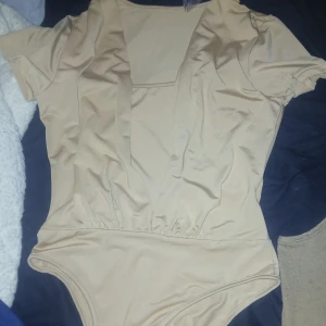 Beige draperad bodysuit med kort ärm - Säljer en beige bodysuit med draperad effekt och v-ringadhalsringning. Bodyn har korta ärmar och är tillverkad i stretchigt material med 95% polyester och 5% elastan. Perfekt att styla med höga byxor eller kjol. Säljer pga satt fel på mig. Möts upp så nära Stockholm södra om möjligt. S-M