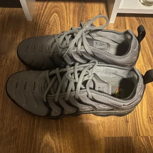 Nike Air Vapormax Plus grå sneakers - Säljer ett par grå Nike Air Vapormax Plus sneakers med svart sula och tydliga Vapormax-detaljer. Skorna har snörning, rund tå och är tillverkade i syntetmaterial. Perfekta för dig som gillar sportig och modern stil.