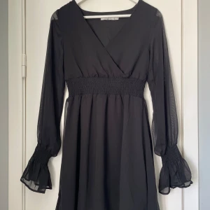 Svart omlottklänning med volangärm - Säljer en svart omlottklänning med långa, transparenta ärmar och smock i midjan. Klänningen har v-ringning och volangdetaljer vid ärmsluten. Perfekt för dig som gillar en klassisk och stilren look.