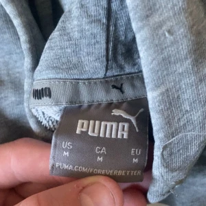 Grå hoodie från Puma - Säljer en grå hoodie från Puma i storlek M. Tröjan har en stor vit Puma-logga framtill, huva med dragsko och en klassisk känguruficka. Perfekt för en avslappnad och sportig stil.