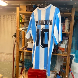”Hand of god” fotbolls Jersey Maradonna.  - Klassisk Argentina fotbollströja med blåvita ränder, Maradona och nummer 10 på ryggen. Broderad AFA-logga på bröstet och Le Coq Sportif-märke. Rund hals och korta ärmar. Perfekt för dig som älskar fotbollshistoria. Köpt på hand of god i Barcelona, exakt kopia av Mardonnas Jersey i vm. 
