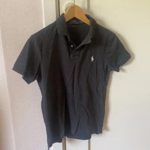 Mörkgrå pikétröja från Polo Ralph Lauren - Säljer en mörkgrå pikétröja från Polo Ralph Lauren med klassisk krage och knappslå. Tröjan har korta ärmar och en broderad logga på bröstet. Perfekt för en stilren och avslappnad look.