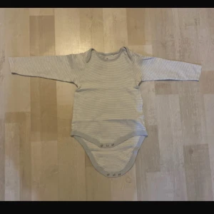 Randig långärmad body - Säljer en söt långärmad body med tunna grå och vita ränder. Bodyn har praktiska knappar i grenen för enkel på- och avklädning. Storlek 92 cm