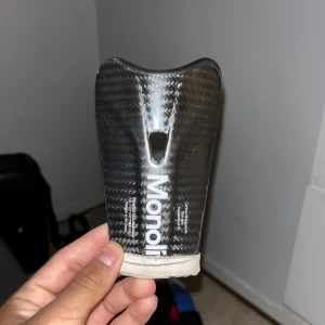 Svart benskydd i kolfiber från Mono - Säljer ett svart benskydd i kolfiber från Mono. Benskyddet har en sportig design med vit logga och ett litet hål i mitten för ventilation. Lätt och smidigt skydd som sitter bra på benet. Perfekt för fotbollsträning eller match.