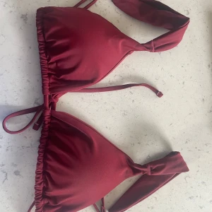 Vinröd bikini med knyt - Säljer en vinröd bikini med trekantsöverdel och knyt i  ryggen. Underdelen har breda band i sidorna och en klassisk tanga-modell. Perfekt för stranden eller poolen!