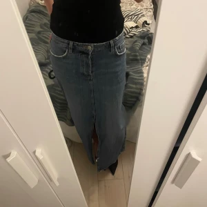 Lång jeanskjol med slits - Säljer en klassisk blå lång jeanskjol med låg midja och en tydlig slits framtill. Kjolen har femficksmodell och stängs med knapp. Perfekt för dig som gillar en tidlös och avslappnad stil.