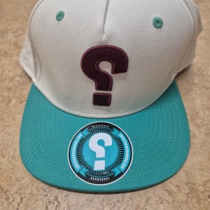 Vit och turkos keps från Sweet Skateboards - Säljer en keps från Sweet Skateboards med vit krona och turkos skärm. Framtill finns ett broderat frågetecken i vinrött. Justerbar snapback baktill. Helt ny, oanvänd.