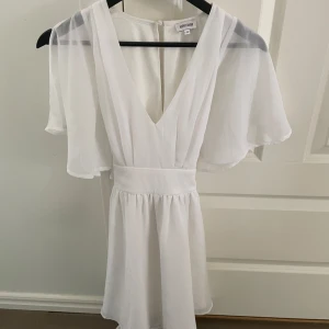 Vit kortklänning med v-ringning från Bubbleroom - Superfin vit klänning från Bubbleroom med djup v-ringning, korta vida ärmar och knytband i midjan. Perfekt för sommarens alla tillfällen eller till studenten! Lätt och luftig modell med markerad midjan fint!