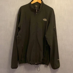 Svart vindjacka från The North Face XXL - Svart vindjacka från The North Face i storlek XXL. Jackan har dragkedja framtill, två sidofickor med dragkedja och justerbar dragsko nedtill. Tillverkad i slitstark polyester och med TNF Apex-tyg för extra skydd mot vind. Perfekt för dig som gillar stilren och funktionell design.