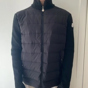 Svart pufferjacka från Moncler - Säljer en svart pufferjacka från Moncler med stickade ärmar och hög krage. Jackan har dragkedja framtill, quiltad front och Moncler-logga på ärmen. Snygg röd Moncler-text längs sidan på ärmen ger en sportig vibe. Perfekt för höst och vinter. Passar för er i L-XL helt ny. 