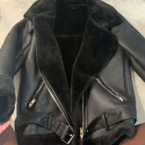 Svart pälsjacka med bälte och dragkedjor - Säljer en svart pälsjacka med bred krage och mjukt foder. Jackan har två sneda dragkedjor framtill, bälte i midjan och detaljer vid ärmslut. Perfekt för dig som vill ha en cool och varm look under kalla dagar.bälte saknas 