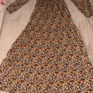 Blommig långklänning med knappar - Långklänning med färgglatt blommönster i gult, blått, rött och vitt på mörk botten. Klänningen har långa ärmar, v-ringning och svarta knappar framtill. Materialet känns lätt och flowy, perfekt för dig som gillar boho-vibes och vill sticka ut.