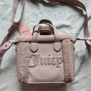 Rosa axelväska från Juicy Couture - Supersöt ljusrosa axelväska från Juicy Couture med fluffiga teddy-detaljer och broderad logga framtill. Väskan har både handtag och justerbar axelrem, perfekt för att bära crossbody. Materialet är en mix av syntet och teddy, vilket ger en trendig och mysig look.
